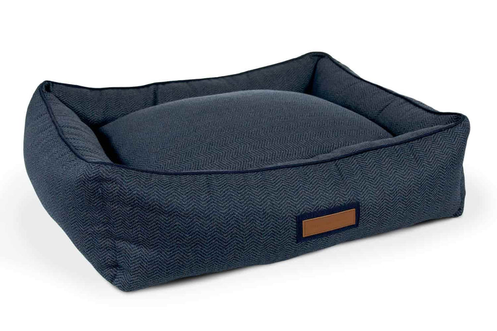 The navy newfie hugger dog bed from the houndry is a simple navy with herringbone patterned texture. The houndry logo is on a leather patch in the bolster's center